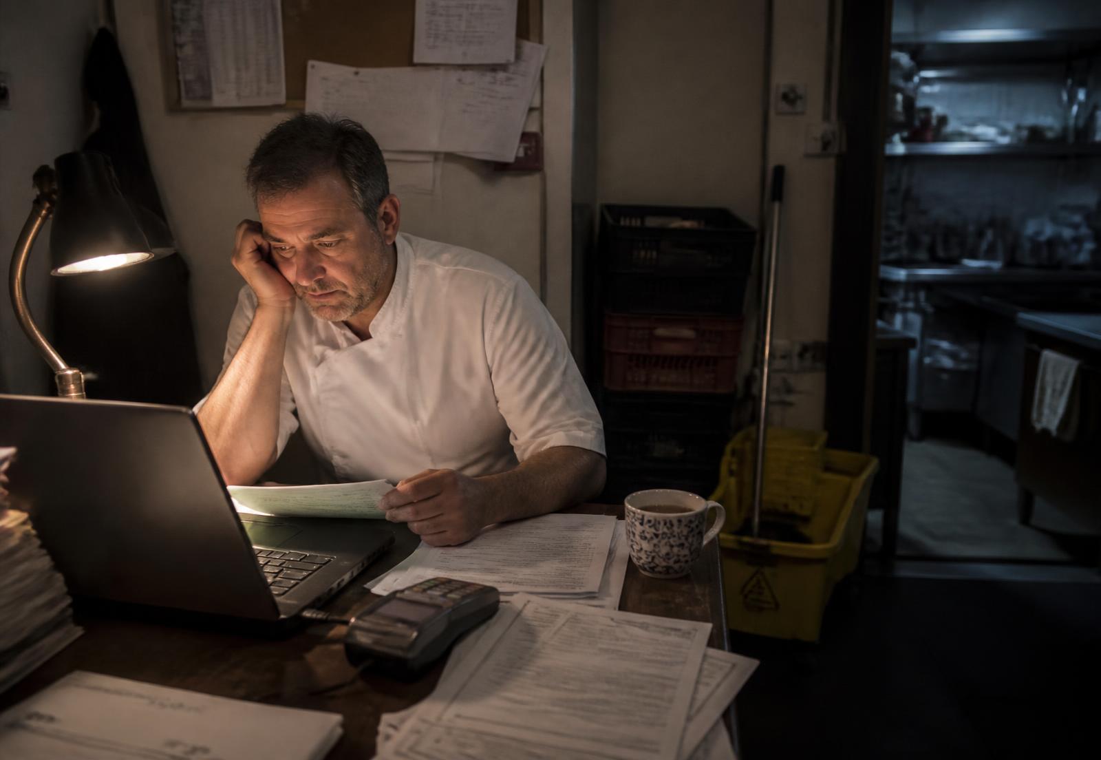 Late at night in a restaurant back office: the owner in chef whites at a desk with a laptop, a card terminal, statements and bills, the dark kitchen visible through the doorway.