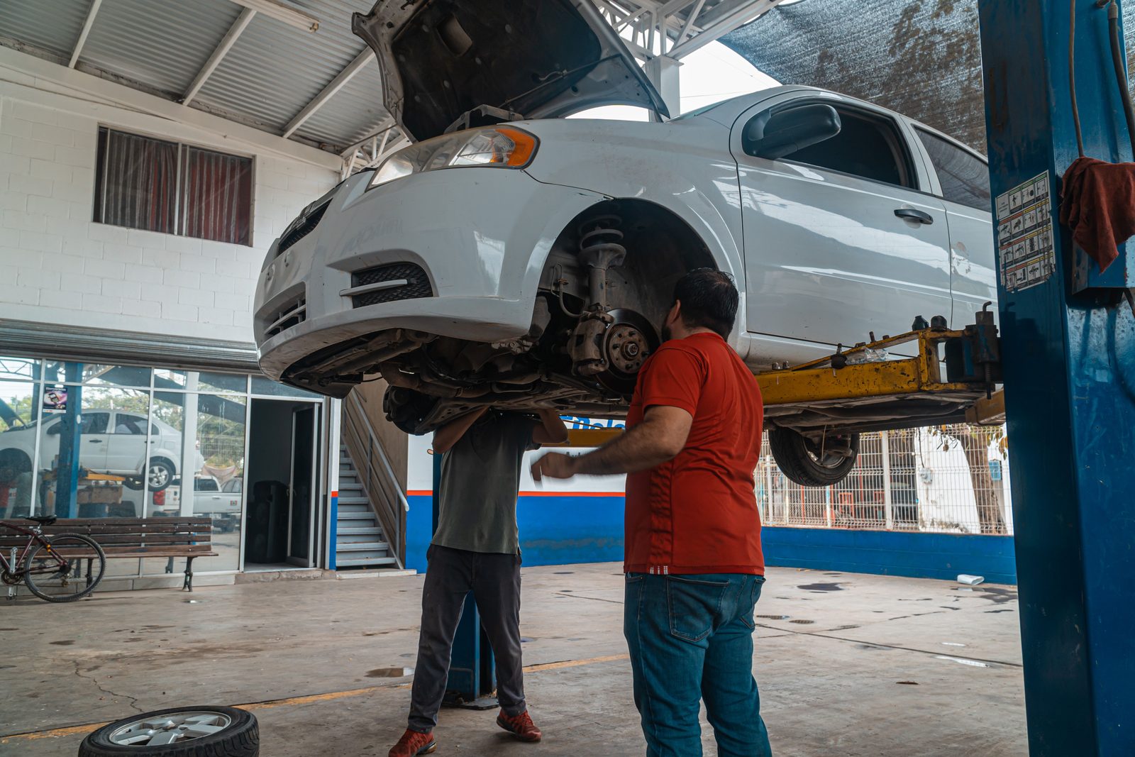Mechanics servicing a car inside a busy independent repair garage, with the vehicle raised on a workshop lift.