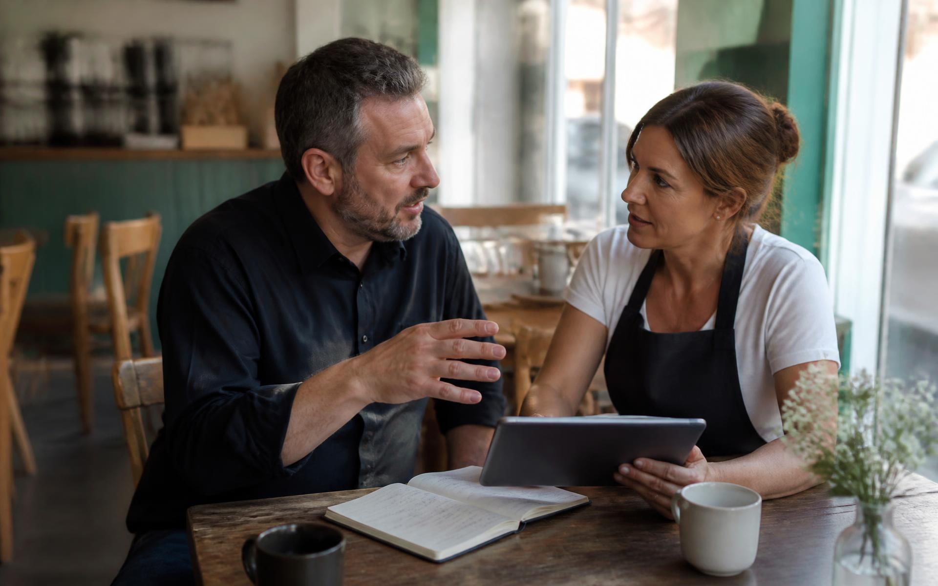 A cafe owner in an apron talking across a table with an adviser, a notebook and tablet between them.